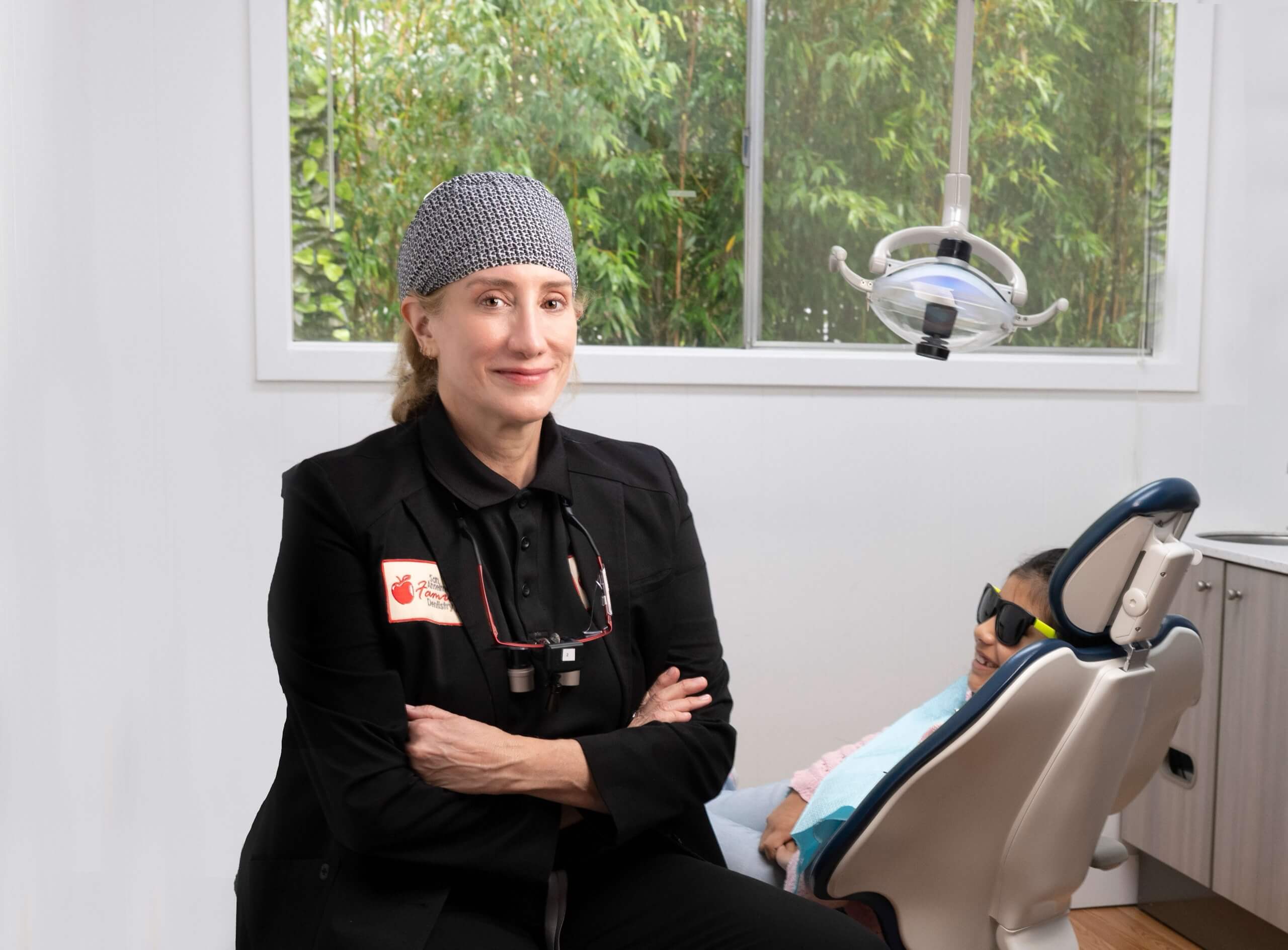 Smiling San Anselmo dentist with arms crossed, beside a relaxed child patient in a dental chair wearing sunglasses. Bright, modern office.