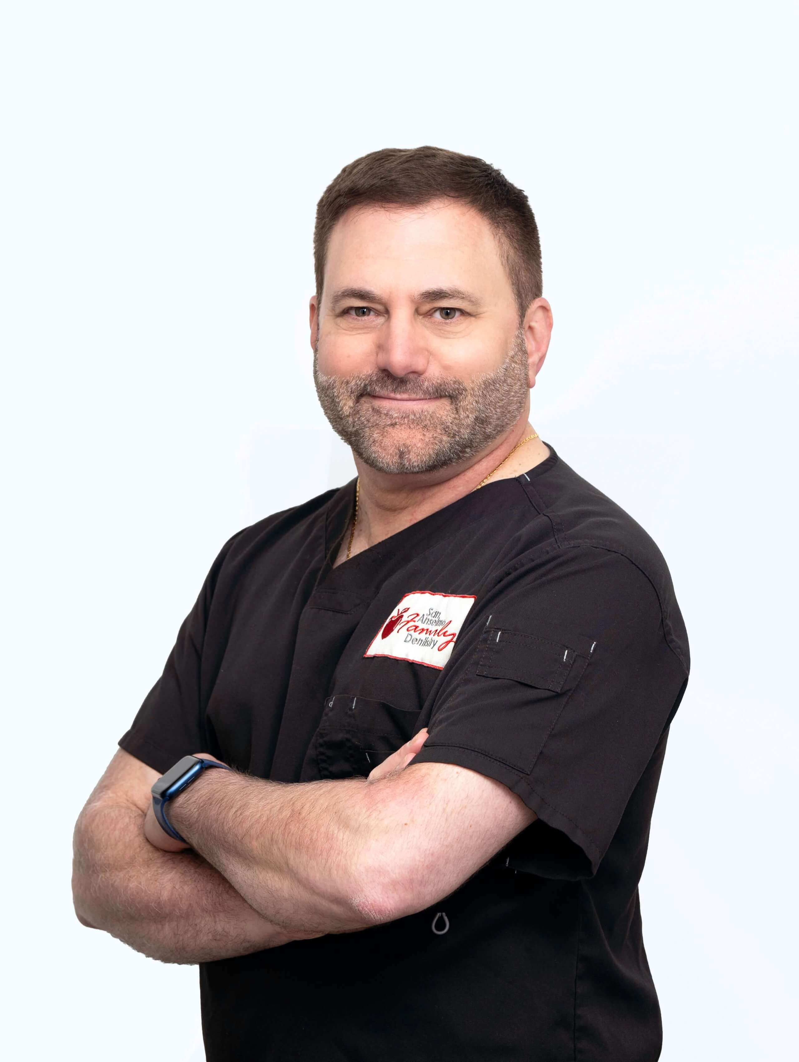 Dr. Anthony Costella, a friendly male dentist with a beard, smiles confidently with arms crossed, wearing black scrubs with the San Anselmo Family Dentistry logo.