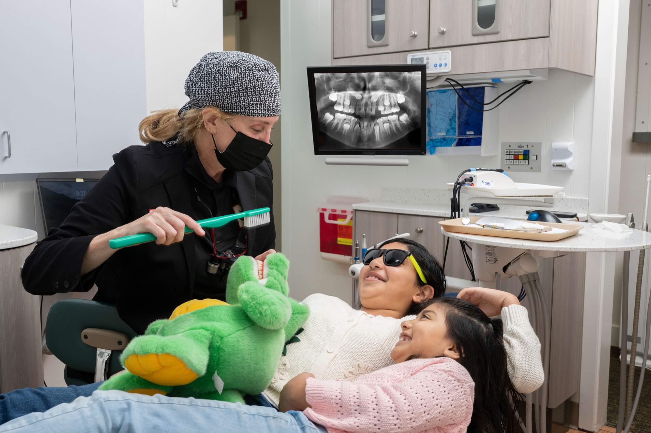 Dental professional teaches two smiling girls about brushing with a large tooth model and plush toy in a modern, family-friendly dental office.