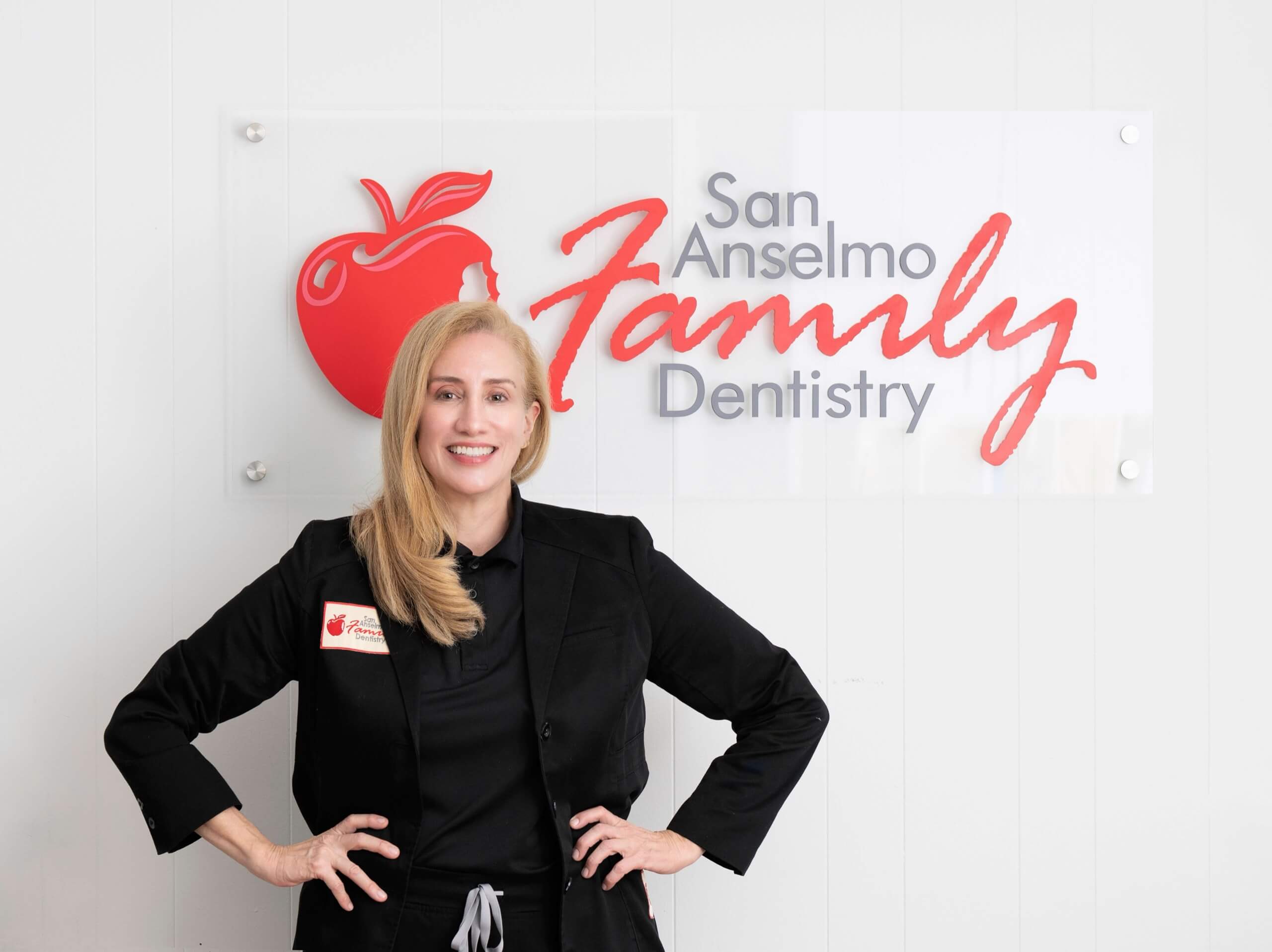 Dr. Rosemarie Goldstein in black scrubs and blazer stands in front of San Anselmo Family Dentistry logo sign on a white wall.