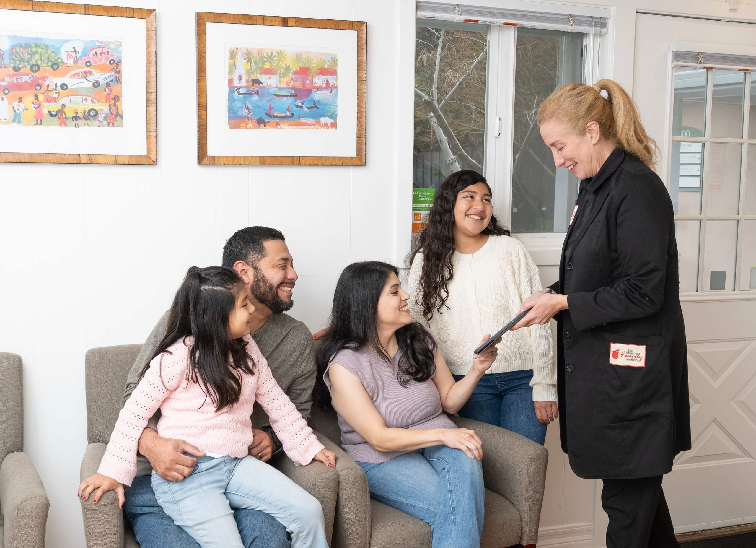 Dental office staff member with a tablet engaging with a smiling family of four in the waiting room at San Anselmo Family Dentistry.